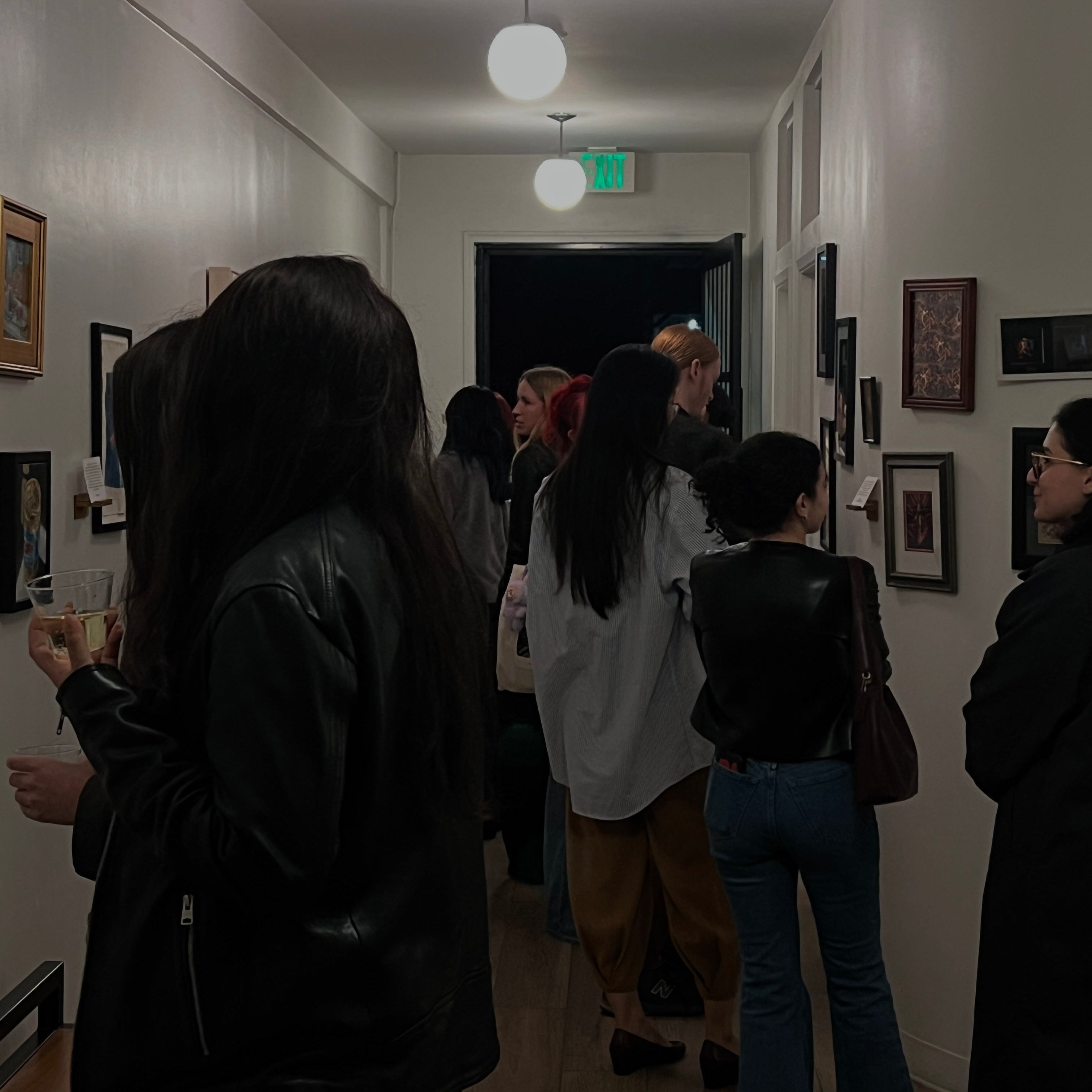 A group of people standing in a dimly lit hallway at Espresso Social Club with framed artwork on the walls, viewed from behind a woman in a black leather jacket.