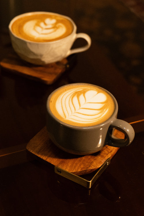 Two cups of latte coffee on wooden coasters, one in a white textured ceramic cup and one in a gray ceramic cup, both featuring leaf-shaped latte art on a dark wooden table.