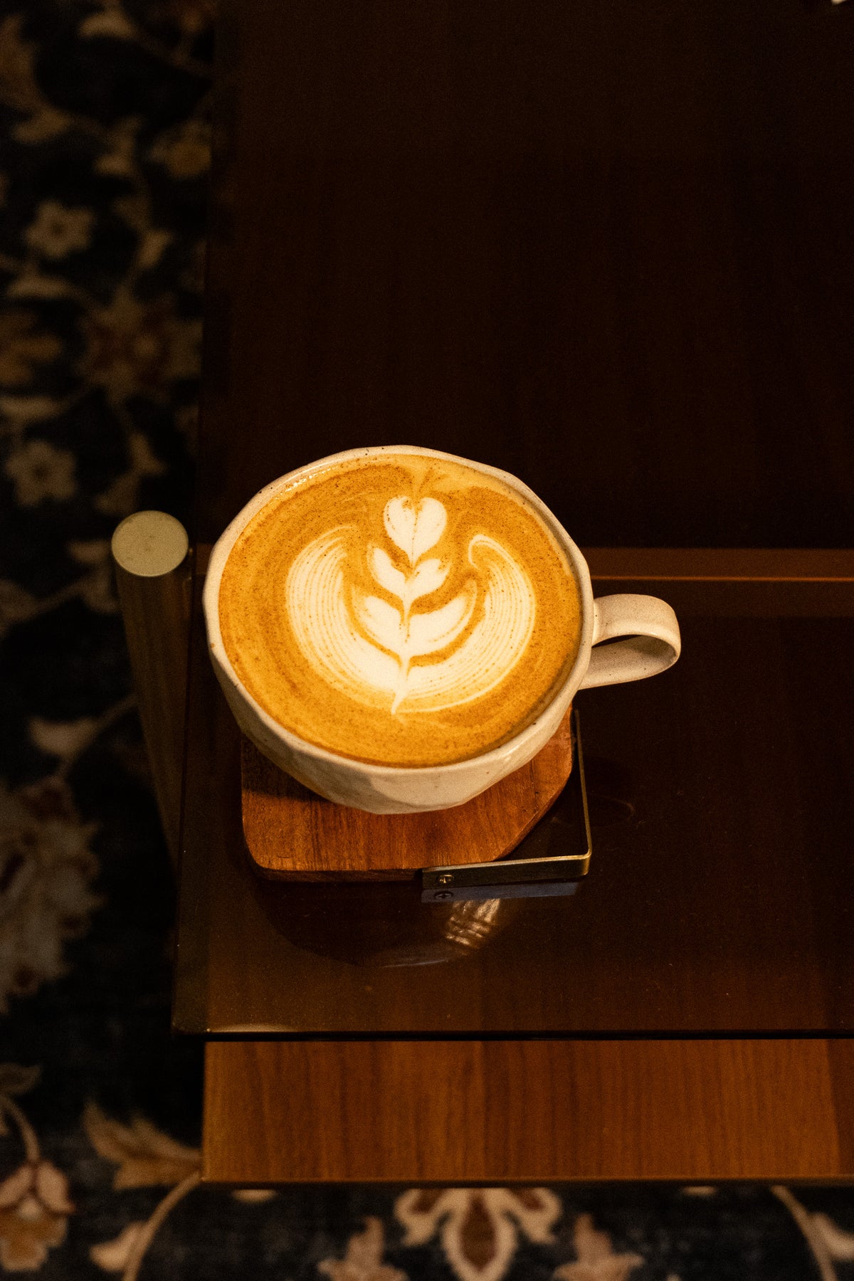 A cup of latte coffee with leaf-shaped latte art sits on a wooden coaster on a dark wooden table.