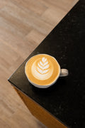 A white ceramic cup of latte coffee with leaf-shaped latte art, resting on a black countertop with a light wood floor in the background.