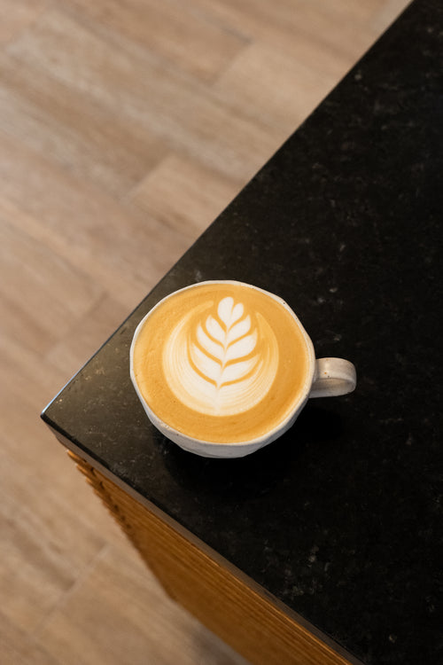 A white ceramic cup of latte coffee with leaf-shaped latte art, resting on a black countertop with a light wood floor in the background.